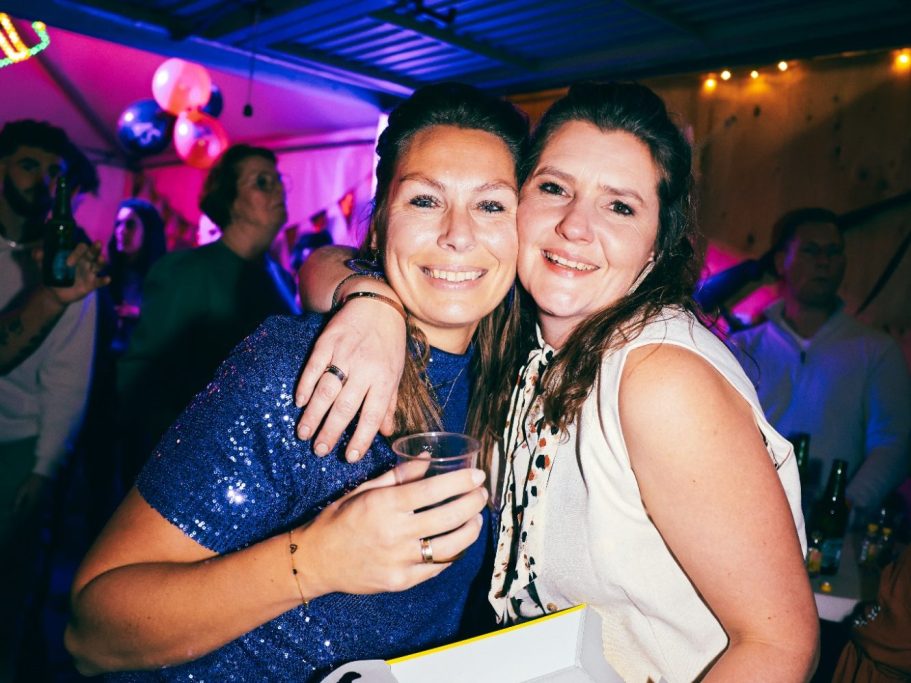 Twee vrouwen lachend met een drankje op een feest met kleurige verlichting.