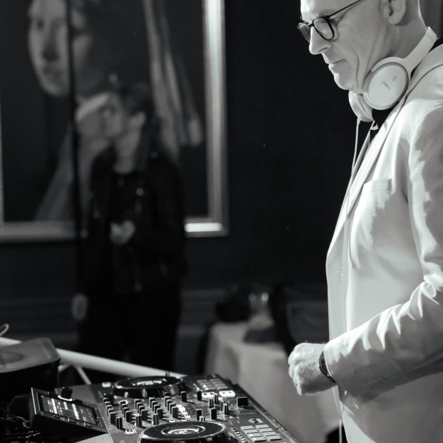 Een DJ in een wit jasje draait muziek voor een publiek in een sfeervolle omgeving.
