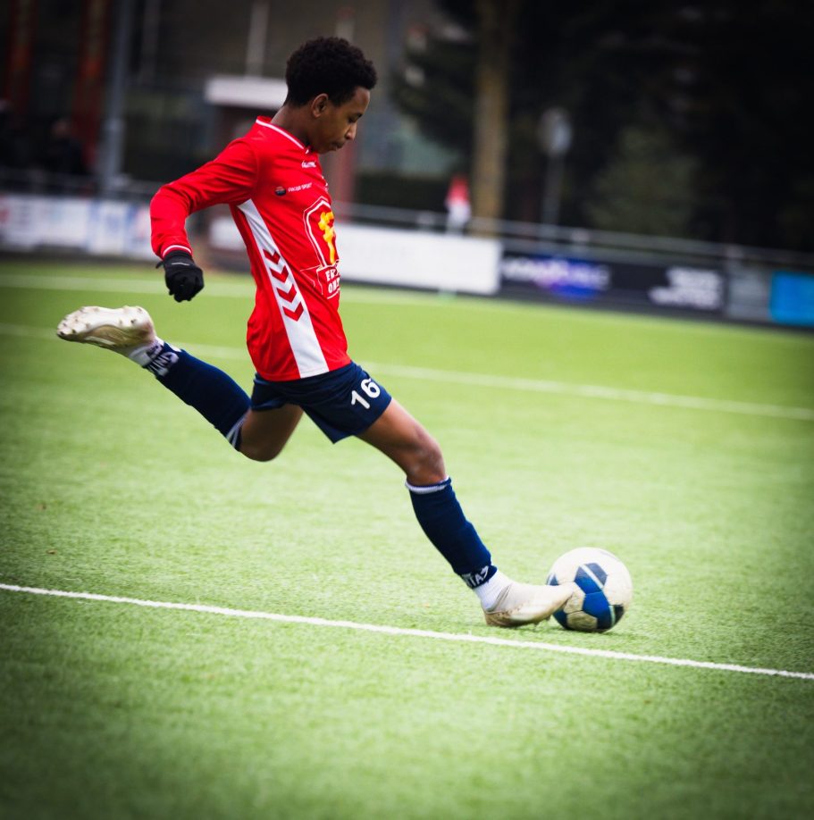 Jongeman in rode voetbalshirt schopt een bal op een grasveld.