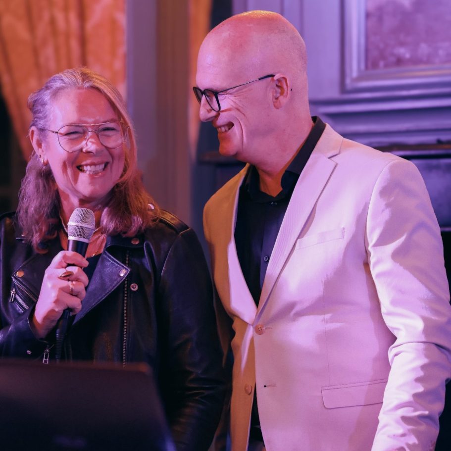 Twee mensen lachen en praten tijdens een presentatie, met een microfoon en een projector.